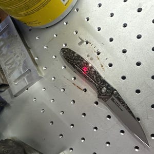 May include: A silver folding knife with an ornate, engraved handle and a sharp blade. The handle features a detailed floral pattern. A red light is visible on the knife. The knife is on a metal surface with evenly spaced holes.