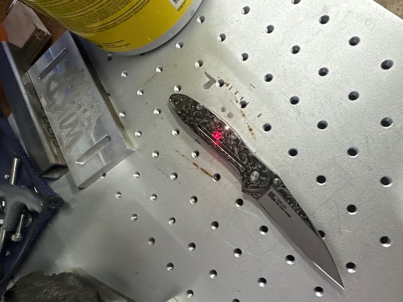 May include: A silver folding knife with an ornate, engraved handle and a sharp blade. The handle features a detailed floral pattern. A red light is visible on the knife. The knife is on a metal surface with evenly spaced holes.