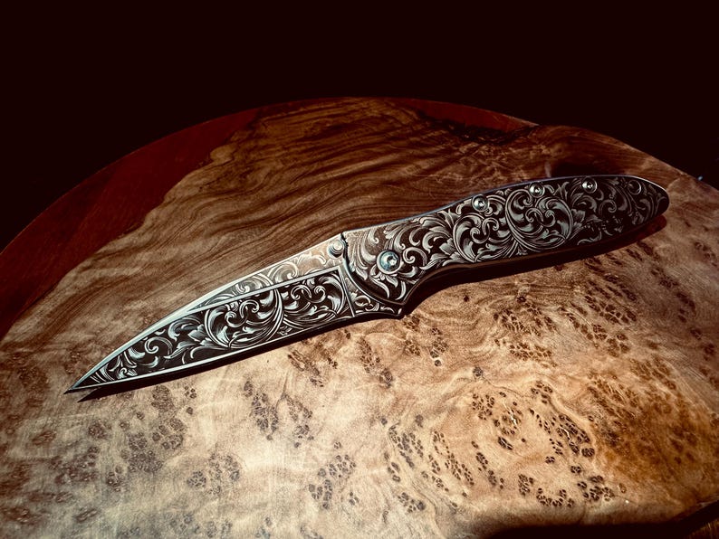 May include: An intricately engraved folding knife with a silver-colored blade and handle. The handle features detailed floral patterns and a small, round, green accent. The knife is displayed on a wooden surface with a natural grain.
