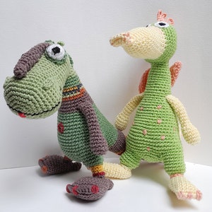 May include: Two crocheted stuffed toys, one green with brown accents and one green with yellow accents. Both toys have large eyes and are standing on a white surface.