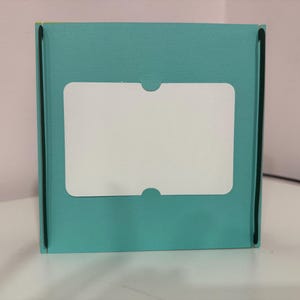 May include: A teal-colored box with a white, rectangular label. The label has rounded corners and a cutout at the top and bottom. The box has black trim along the edges.