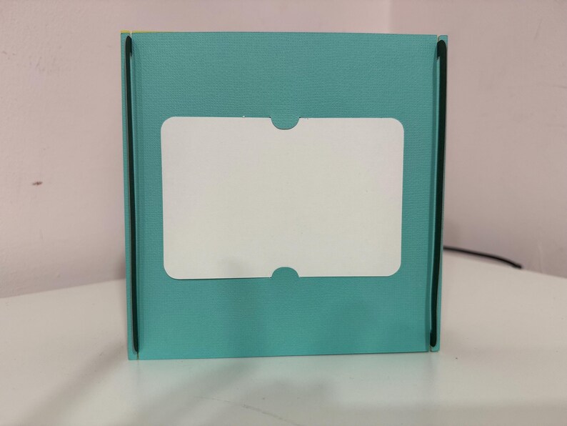 May include: A teal-colored box with a white, rectangular label. The label has rounded corners and a cutout at the top and bottom. The box has black trim along the edges.