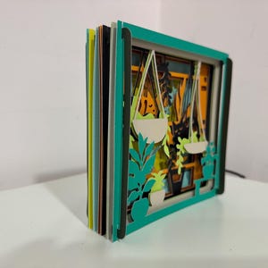 May include: A layered paper art piece featuring a window scene with hanging plants. The artwork has a teal frame and multiple layers of colorful paper, creating a 3D effect. The scene includes white hanging planters and green foliage.