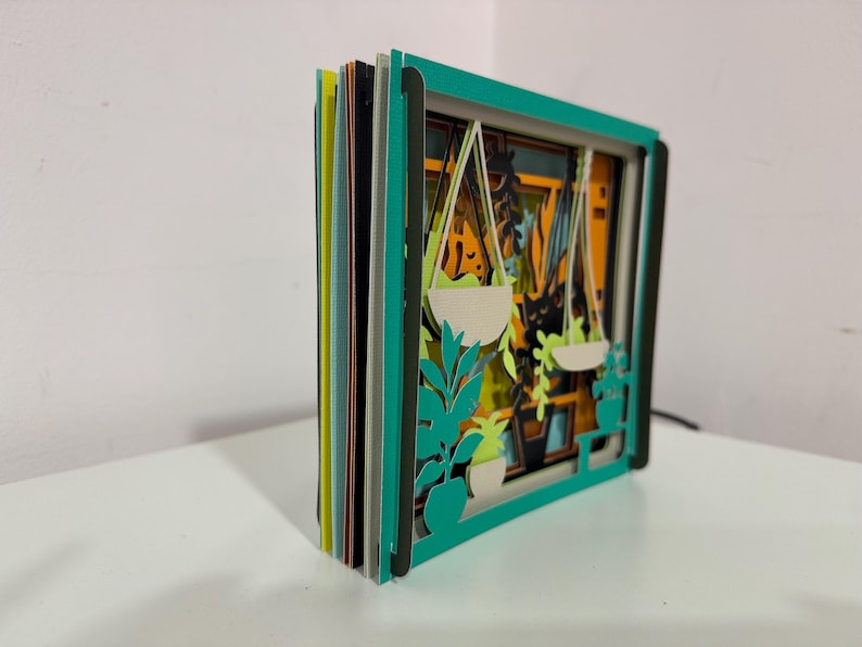 May include: A layered paper art piece featuring a window scene with hanging plants. The artwork has a teal frame and multiple layers of colorful paper, creating a 3D effect. The scene includes white hanging planters and green foliage.