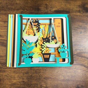 May include: A layered paper art piece featuring a window scene with hanging plants, potted plants, and a cat silhouette. The artwork uses shades of green, orange, yellow, and black. The piece is set against a wooden background.