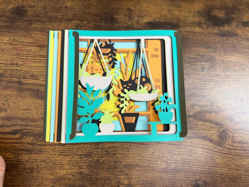 May include: A layered paper art piece featuring a window scene with hanging plants, potted plants, and a cat silhouette. The artwork uses shades of green, orange, yellow, and black. The piece is set against a wooden background.