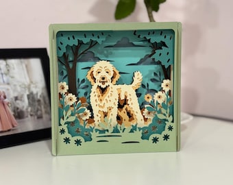 May include: A layered paper art piece featuring a dog in a natural setting. The artwork is housed in a square, light green frame. The dog is light brown with a fluffy coat, surrounded by trees, flowers, and foliage. The background is a light blue.