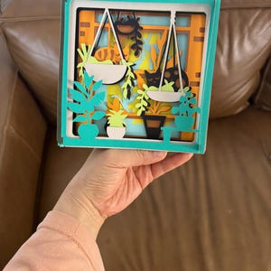 May include: A layered paper art piece depicting a window scene with hanging plants, a cat, and potted greenery. The artwork features a teal frame and a vibrant color palette of orange, green, and black. The piece is held by a person.
