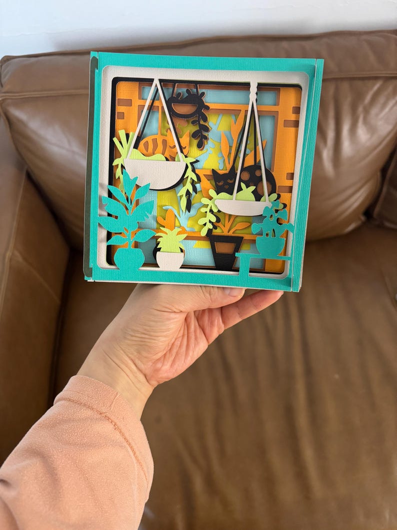 May include: A layered paper art piece depicting a window scene with hanging plants, a cat, and potted greenery. The artwork features a teal frame and a vibrant color palette of orange, green, and black. The piece is held by a person.