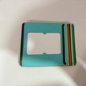 May include: A stack of colorful file folders. The top folder is a bright teal with a white rectangular window. The edges are black. Other folders in the stack are orange, green, and yellow.