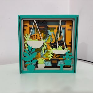May include: A layered paper art piece in a teal frame. The artwork depicts a window scene with hanging plants and cats. The background is orange, with white hanging planters and green foliage. Black cat silhouettes are visible.