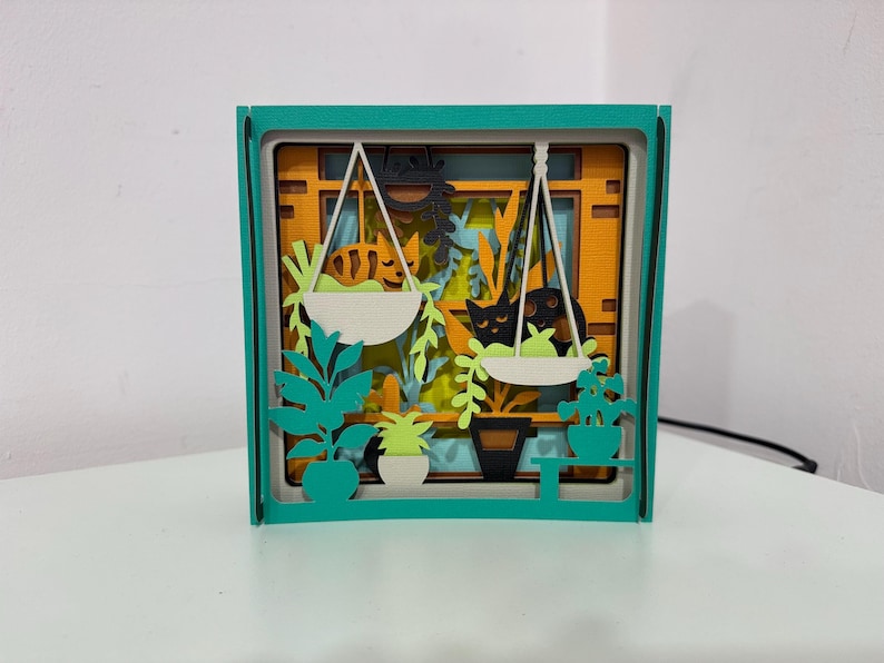 May include: A layered paper art piece in a teal frame. The artwork depicts a window scene with hanging plants and cats. The background is orange, with white hanging planters and green foliage. Black cat silhouettes are visible.