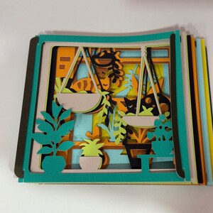 May include: A layered paper art piece depicting a window scene with hanging plants, potted plants, and a cat. The artwork features teal, orange, yellow, and black colors. The piece is a stack of cut paper layers.