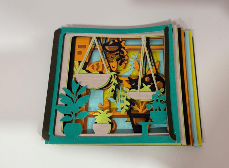 May include: A layered paper art piece depicting a window scene with hanging plants, potted plants, and a cat. The artwork features teal, orange, yellow, and black colors. The piece is a stack of cut paper layers.