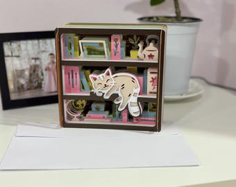 May include: A 3D paper art bookshelf with a sleeping cat. The shelves are filled with books, a teapot, and other decorative items. The color palette includes shades of brown, pink, and green. A white envelope is in front of the bookshelf.