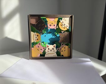 May include: A layered paper art piece featuring a square frame with a scene of cartoon cats. The cats are in various colors, including pink, yellow, and brown, and are arranged around a blue and green background.