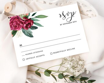 May include: White RSVP card with burgundy floral watercolor design. The card includes the text "RSVP by November 20th" and checkboxes for "Joyfully Accepts" and "Regretfully Decline". The card is decorated with green leaves and white flowers.