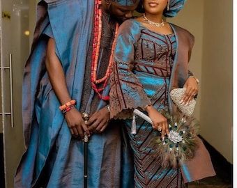 May include: A couple in traditional Nigerian attire. The man wears a blue robe and cap, holding a cane. The woman wears a matching blue and brown dress, head wrap, and holds a fan. They are adorned with coral beads.