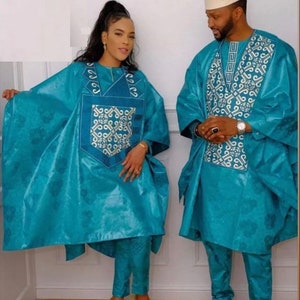 May include: Matching teal outfits. The ensemble includes a tunic with embroidered details and matching pants. The man wears a white cap and black shoes. The woman wears gold heels. The outfits have a shiny, textured appearance.