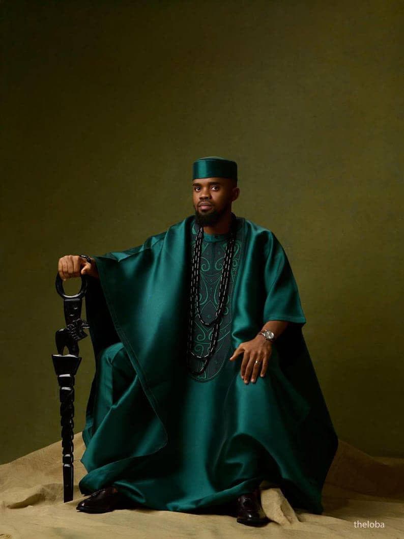 May include: A person wearing a teal green traditional outfit, including a tunic, pants, and a matching cap. The outfit features intricate embroidery and is accessorized with a long beaded necklace and a decorative staff. The background is a solid olive green.