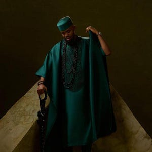 May include: A teal green traditional outfit, including a tunic, hat, and cape-like garment. The tunic features embroidered detailing. The person is holding a black cane. The background is a dark, neutral color.
