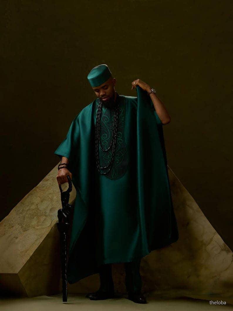 May include: A teal green traditional outfit, including a tunic, hat, and cape-like garment. The tunic features embroidered detailing. The person is holding a black cane. The background is a dark, neutral color.