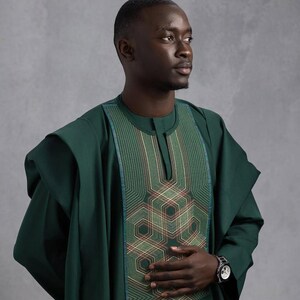 May include: A dark green traditional African outfit with intricate geometric embroidery on the chest. The outfit features a round neckline and wide sleeves. A silver wristwatch is visible on the wrist.