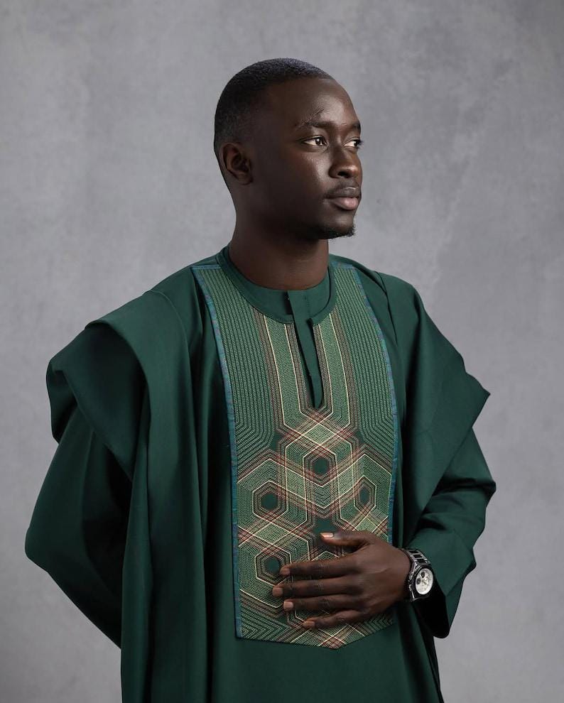 May include: A dark green traditional African outfit with intricate geometric embroidery on the chest. The outfit features a round neckline and wide sleeves. A silver wristwatch is visible on the wrist.