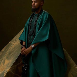 May include: A person wearing a teal green traditional outfit, including a matching hat and a flowing robe. The outfit is paired with a black beaded necklace and a silver wristwatch. The background is a dark, textured surface.