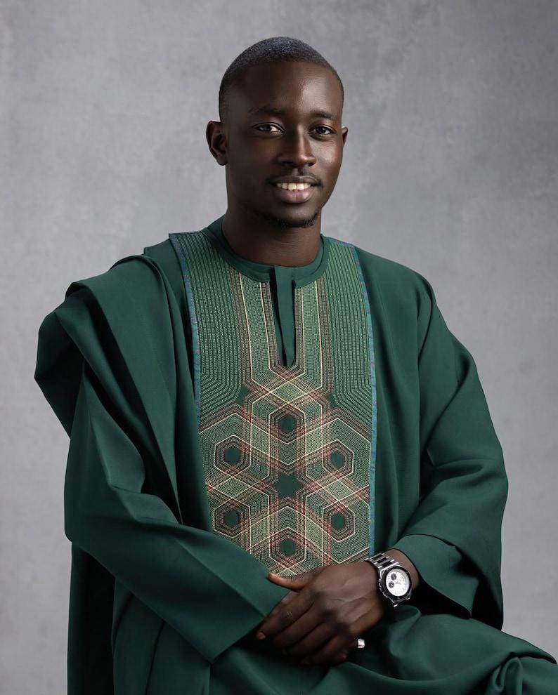 May include: A dark green traditional outfit with intricate embroidery on the chest. The outfit includes a long-sleeved tunic and matching trousers. A silver wristwatch with a black face is visible on the wrist.