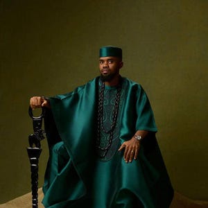 May include: A person wearing a teal green traditional outfit, including a tunic, pants, and a matching cap. The outfit features intricate embroidery and is accessorized with a long beaded necklace and a decorative staff. The background is a solid olive green.