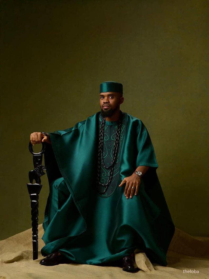 May include: A person wearing a teal green traditional outfit, including a tunic, pants, and a matching cap. The outfit features intricate embroidery and is accessorized with a long beaded necklace and a decorative staff. The background is a solid olive green.