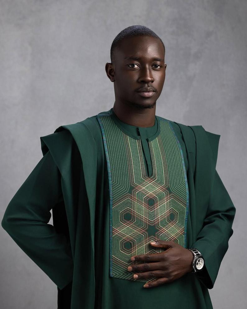 May include: A dark green traditional outfit with intricate geometric embroidery on the chest. The outfit includes a long-sleeved tunic and matching draped shoulder pieces. A silver wristwatch is visible on the wrist.