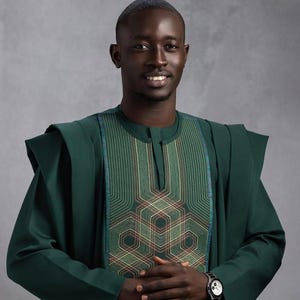 May include: A man wearing a dark green traditional outfit. The outfit features a detailed geometric pattern on the chest and sleeves. A black wristwatch is visible on the left wrist. The background is a solid gray.