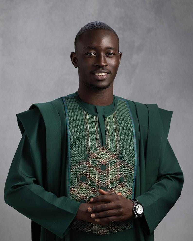 May include: A man wearing a dark green traditional outfit. The outfit features a detailed geometric pattern on the chest and sleeves. A black wristwatch is visible on the left wrist. The background is a solid gray.