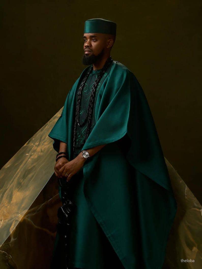 May include: A person wearing a teal green traditional outfit, including a matching hat and cape-like garment. The outfit is paired with a black beaded necklace and a silver wristwatch. The background is a dark, textured surface.