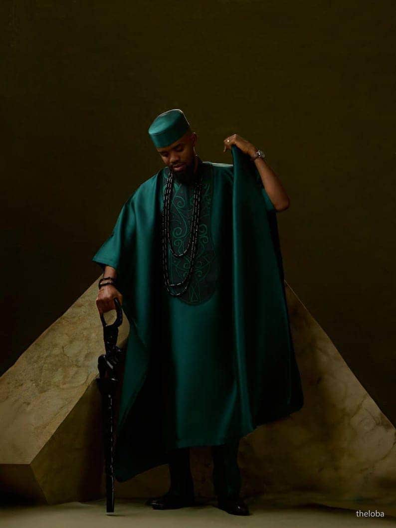 May include: A teal green traditional outfit, including a tunic, hat, and cape-like garment. The tunic features embroidered detailing around the neckline. The person is holding a black cane. The background is a dark, neutral color.
