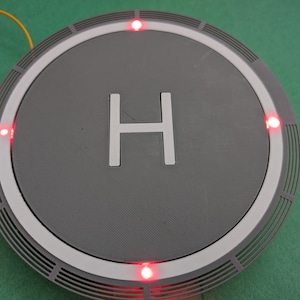 May include: A grey circular platform with a white ring and a white letter "H" in the centre. Four red lights are visible around the edge of the platform.