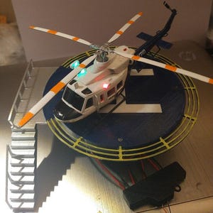 May include: A white and black toy helicopter with orange and white stripes on the blades. The helicopter is sitting on a blue and yellow circular platform with a white letter 'L' on it. The helicopter has red and green lights on it.