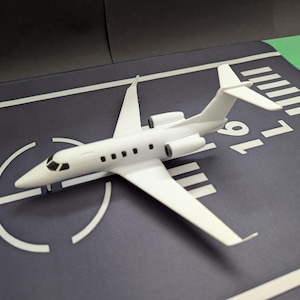 Può includere: Un modello di aereo bianco, simile a un jet privato, è posizionato su una superficie blu scuro con segnaletica bianca della pista. Il velivolo in miniatura ha ali, coda e finestre dettagliate. La pista ha segni e numeri bianchi.