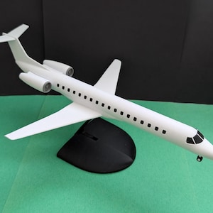 Può includere: Un modello di aereo bianco con due motori, ali e un timone di coda. Il modello è montato su un supporto nero. L'aereo ha una fila di piccole finestre lungo la fusoliera.