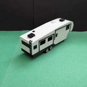 May include: A white and black toy travel trailer with two axles and four wheels. The trailer has a rectangular shape with black accents around the windows and doors. Two black rectangular features are on the roof. The trailer is on a green surface.