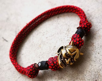 Puede incluir: Un brazalete de paracord rojo y negro con un dije de máscara de demonio dorado.