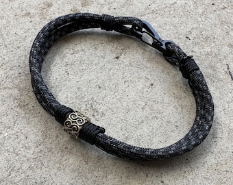 May include: A black and grey rope necklace with a silver metal bead. The bead has a decorative swirl pattern.