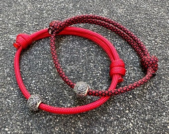 May include: Two red rope bracelets with black accents and silver beads. The bracelets are tied together in a knot.