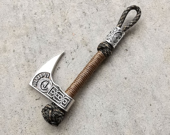 May include: A silver axe keychain with a brown and green paracord handle. The axe head features a compass design and runes around the edge.