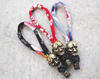 May include: Three gold-colored keychains with a Japanese demon mask design. Each keychain has a different patterned fabric cord: red with white flowers, blue with white flowers, and black with white flowers.