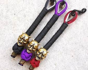 May include: Three keychains with gold demon mask charms. The keychains are made with black, purple, and red paracord. Each keychain has a black metal hook.