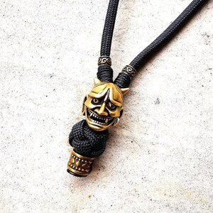 May include: A black paracord necklace with a gold-colored oni mask bead and two decorative metal beads.