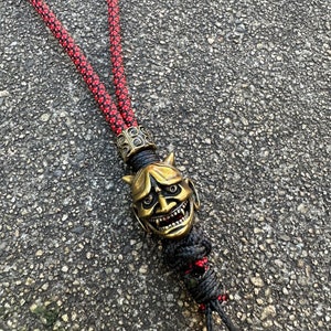 May include: A red and black paracord lanyard with a gold-coloured oni mask charm. The mask has a fierce expression with horns, sharp teeth, and red eyes.